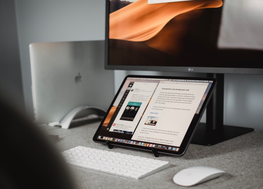 A home office setup with a tablet displaying a document in the foreground, a keyboard and mouse in front of it, and a laptop and external monitor in the background.