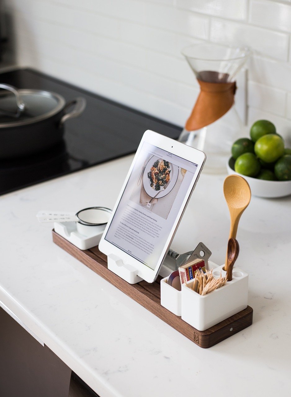 An iPad displaying a recipe sits on a countertop stand in a modern kitchen. The stand also holds containers for cooking utensils, toothpicks, and matches. In the background, a stovetop, a bowl of limes, and a coffee pour-over stand are visible.